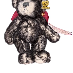 May include: A plush teddy bear with a black and white speckled fur pattern. The bear has a red bow, a small hat, and a tag from "Gund Bears & Co.". The bear is standing on a white background.