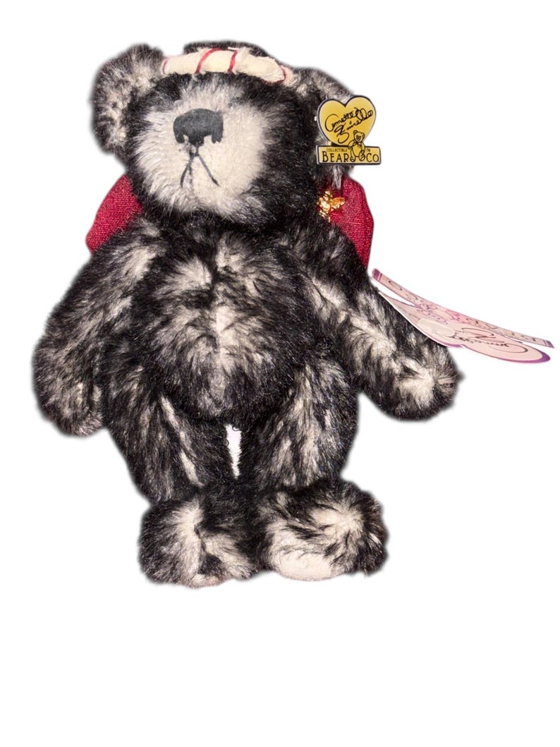 May include: A plush teddy bear with a black and white speckled fur pattern. The bear has a red bow, a small hat, and a tag from "Gund Bears & Co.". The bear is standing on a white background.