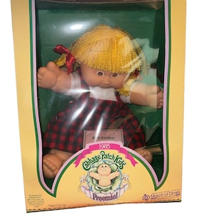 May include: A Cabbage Patch Kids Preemie doll with blonde hair, a red and black plaid dress, and a red bow. The doll is in a box with a 1985 birth certificate and the words "Cabbage Patch Kids Preemie" on it. The box also has the March of Dimes logo.