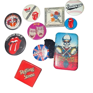 May include: A collection of vintage pinback buttons and a rectangular magnet. The buttons feature band logos like The Rolling Stones, Motley Crue, and The Kinks. Other buttons display a baseball and a skull graphic. The magnet reads "Rolling Stone".