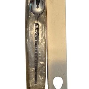 May include: A silver spoon with a decorative handle, presented in its original packaging. The spoon's bowl reflects the surrounding environment. The handle features an ornate floral design. The packaging is a long, rectangular box.