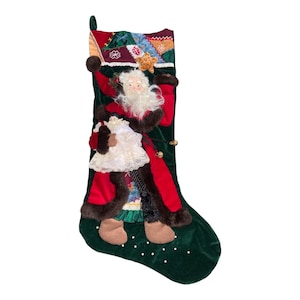 May include: A dark green velvet Christmas stocking featuring a three-dimensional Santa Claus figure wearing a red coat with brown faux fur trim. Santa holds a small white lace-trimmed doll. The cuff has a multicolored patchwork design.