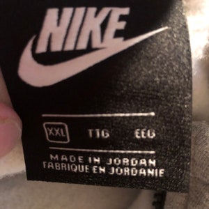 Pode incluir: Uma etiqueta Nike preta e branca com a palavra "NIKE" em letras brancas. A etiqueta tamb&eacute;m inclui o tamanho "XXL", "TTG" e "EEC" em letras brancas. A etiqueta diz "Made in Jordan Fabrique en Jordanie".