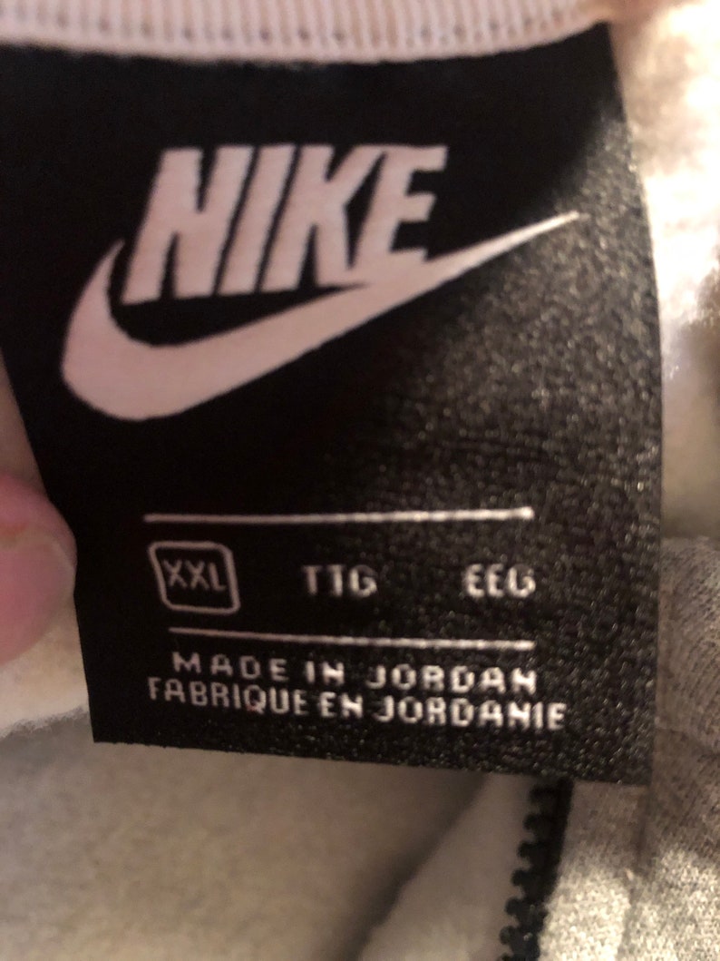 Pode incluir: Uma etiqueta Nike preta e branca com a palavra "NIKE" em letras brancas. A etiqueta tamb&eacute;m inclui o tamanho "XXL", "TTG" e "EEC" em letras brancas. A etiqueta diz "Made in Jordan Fabrique en Jordanie".