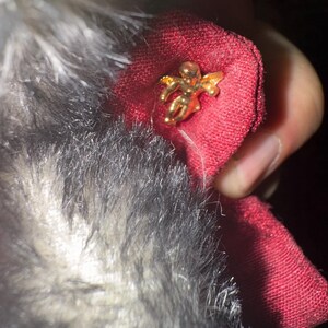 May include: A gold-colored cherub pin is attached to a piece of red fabric. The cherub has outstretched wings and is in a flying pose. The fabric is held in a person's hand, with gray fur in the background.