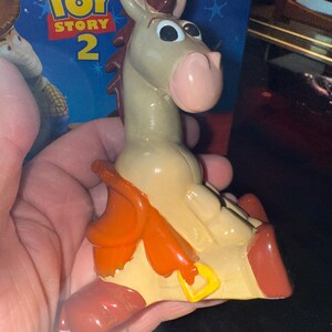 May include: A figurine of Bullseye from Toy Story 2. The toy horse is light tan with a pink nose, brown mane, and red hooves. It has an orange saddle and a yellow buckle. The Toy Story 2 box is in the background.