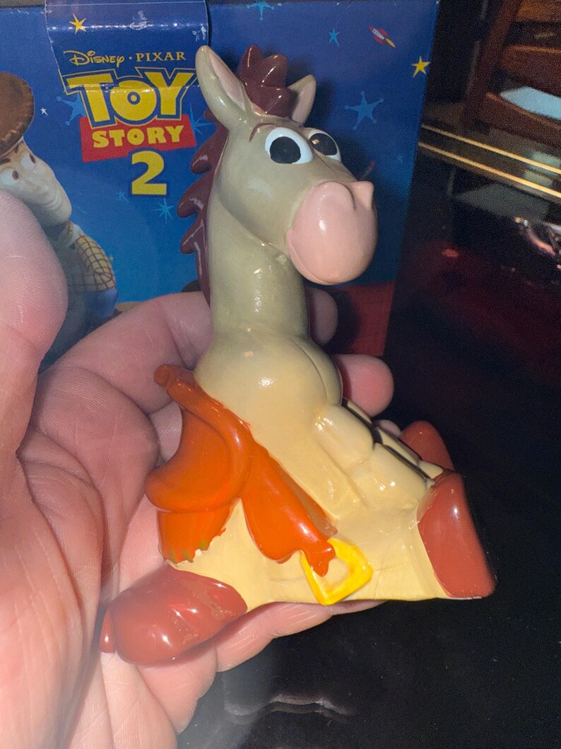May include: A figurine of Bullseye from Toy Story 2. The toy horse is light tan with a pink nose, brown mane, and red hooves. It has an orange saddle and a yellow buckle. The Toy Story 2 box is in the background.