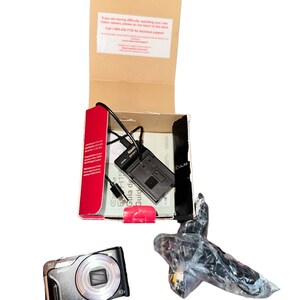 May include: A silver and black digital camera with a 10x zoom lens. The camera is in its original box with a charger and instruction manual. The box is brown with red and white text.