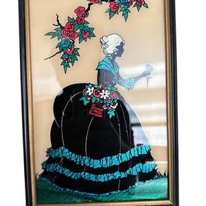 May include: Framed silhouette art featuring a woman in a black dress with blue accents, holding a flower. The artwork includes a floral branch with red and blue flowers. The frame is black with a gold inner edge, creating a vintage aesthetic.
