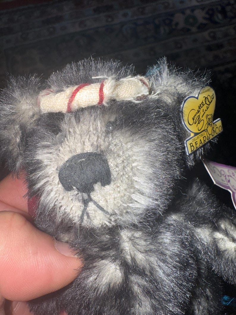 May include: Close-up of a plush teddy bear with gray and black fur. The bear has a black nose and a cream-colored headband with red stripes. A yellow tag with the text "Gommette Bear Co" is attached.