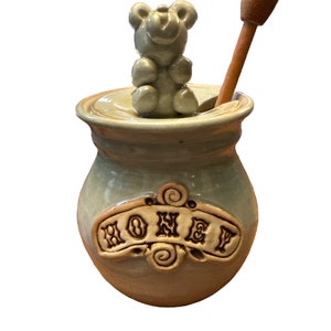 May include: A ceramic honey pot with a bear-shaped lid and a wooden honey dipper. The pot is glazed in a light blue color with a brown accent and the word "HONEY" is written on the front.