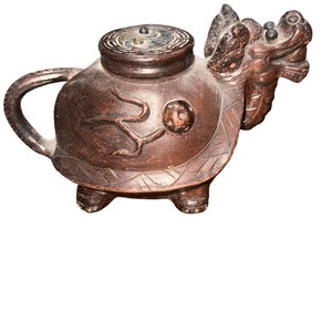 May include: A brown, intricately carved teapot with a dragon head spout and a turtle-like body. The lid is circular with a central knob. The teapot has a handle and is raised on three small feet. The wood has a rich, dark color.
