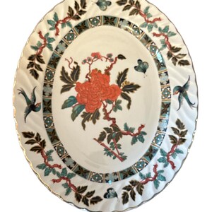 May include: A white oval ceramic plate with a gold rim and a floral design. The design features a red flower with green leaves and branches, and two blue butterflies. The plate has a scalloped edge.