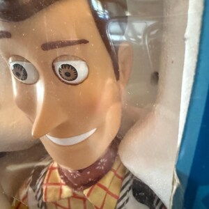 Puede incluir: Primer plano de una mu&ntilde;eca Woody de la franquicia Toy Story. La mu&ntilde;eca tiene una cara bronceada, cabello casta&ntilde;o y una bandana roja. Lleva una camisa a cuadros amarilla, un chaleco negro y una placa de sheriff.