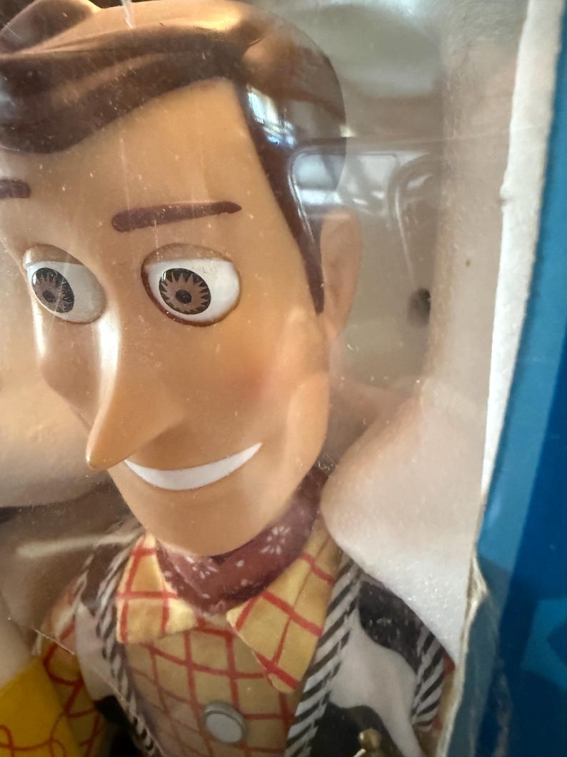 Puede incluir: Primer plano de una mu&ntilde;eca Woody de la franquicia Toy Story. La mu&ntilde;eca tiene una cara bronceada, cabello casta&ntilde;o y una bandana roja. Lleva una camisa a cuadros amarilla, un chaleco negro y una placa de sheriff.