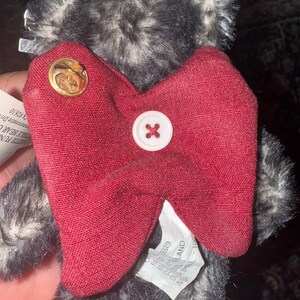 May include: A plush teddy bear with gray and black fur, a red bow tie, and a white button. The bear has a gold pin on the bow tie. The tag says "VETTE FUN" and "STIBLE BEAR C".