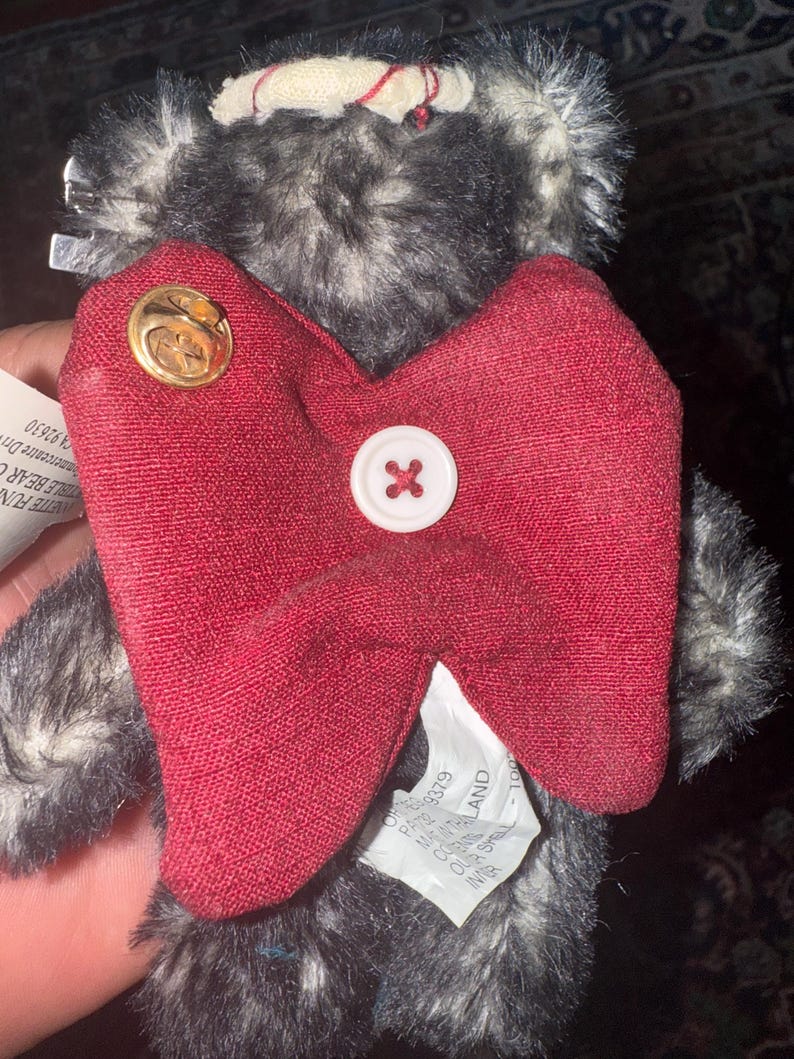 May include: A plush teddy bear with gray and black fur, a red bow tie, and a white button. The bear has a gold pin on the bow tie. The tag says "VETTE FUN" and "STIBLE BEAR C".