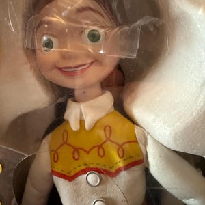 Puede incluir: Una mu&ntilde;eca Jessie de la franquicia Toy Story, empaquetada en un recipiente de pl&aacute;stico transparente. La mu&ntilde;eca tiene una expresi&oacute;n sonriente, cabello rojo y un chaleco amarillo con ribetes rojos y costuras decorativas. La mu&ntilde;eca lleva una camisa blanca con cuello y vaqueros azules.