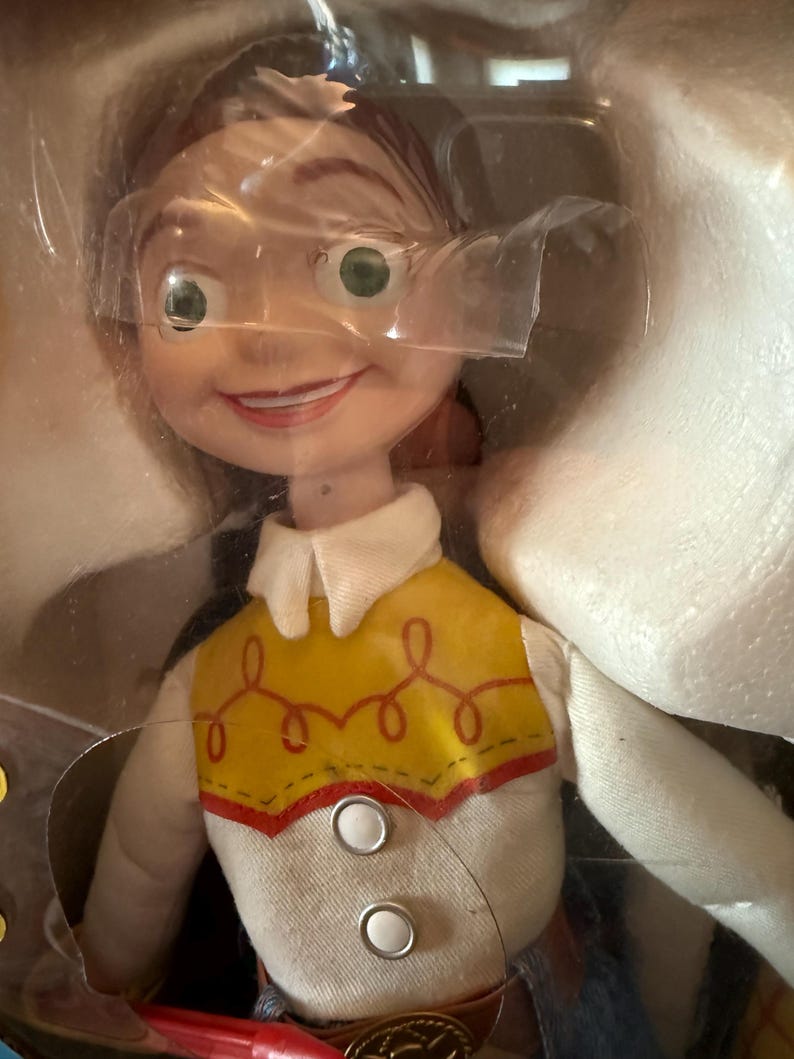 Puede incluir: Una mu&ntilde;eca Jessie de la franquicia Toy Story, empaquetada en un recipiente de pl&aacute;stico transparente. La mu&ntilde;eca tiene una expresi&oacute;n sonriente, cabello rojo y un chaleco amarillo con ribetes rojos y costuras decorativas. La mu&ntilde;eca lleva una camisa blanca con cuello y vaqueros azules.
