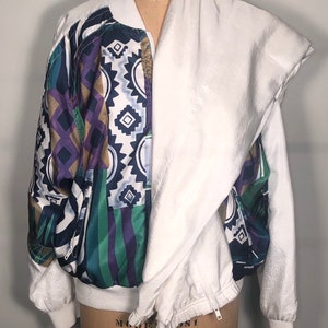 May include: A white bomber jacket with a colorful geometric pattern on one side and a solid white side. The jacket has a zipper closure and a drawstring at the bottom.