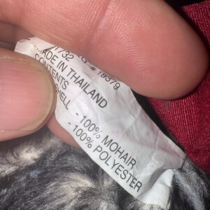 May include: A close-up of a white tag with black text. The tag reads "MADE IN THAILAND" and lists the contents as "100% MOHAIR" and "100% POLYESTER". The tag is held by a person.