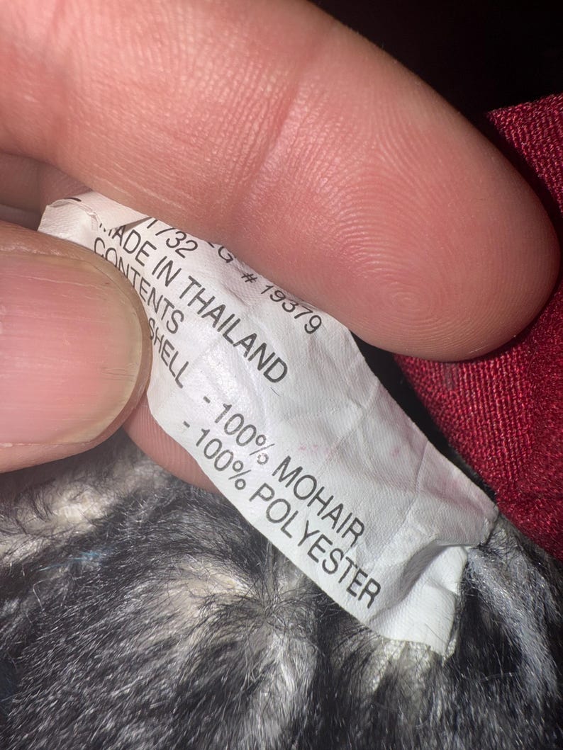 May include: A close-up of a white tag with black text. The tag reads "MADE IN THAILAND" and lists the contents as "100% MOHAIR" and "100% POLYESTER". The tag is held by a person.