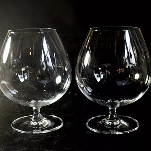May include: Two clear glass brandy snifters with a round bowl and a short stem. The snifters are on a black background.