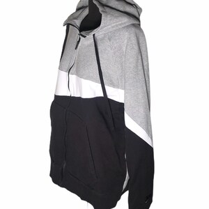 Pode incluir: Uma camisola de capuz cinzenta, branca e preta com fecho de correr. A camisola tem um design de blocos de cor e um log&oacute;tipo da Nike na manga.