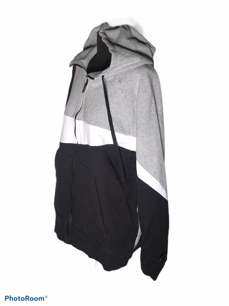 Pode incluir: Uma camisola de capuz cinzenta, branca e preta com fecho de correr. A camisola tem um design de blocos de cor e um log&oacute;tipo da Nike na manga.