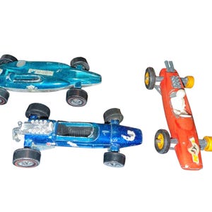 May include: Three vintage toy race cars in teal, blue, and red. The teal car has the number 68 on it. The blue car has a silver engine detail. The red car has the number 57 on it and a white driver figure.