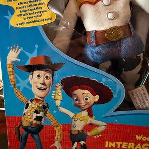 Puede incluir: Un juguete parlante interactivo de Woody y Jessie en caja. El embalaje presenta ilustraciones de los personajes. El texto en la caja dice "&iexcl;PRU&Eacute;BANOS!" y describe las funciones interactivas del juguete. La mu&ntilde;eca Woody lleva una camisa amarilla con estampado de vaca.