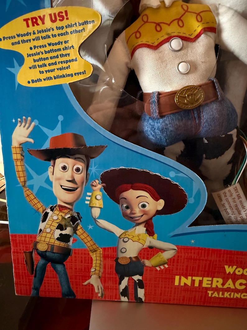 Puede incluir: Un juguete parlante interactivo de Woody y Jessie en caja. El embalaje presenta ilustraciones de los personajes. El texto en la caja dice "&iexcl;PRU&Eacute;BANOS!" y describe las funciones interactivas del juguete. La mu&ntilde;eca Woody lleva una camisa amarilla con estampado de vaca.