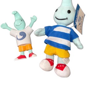 May include: Two turquoise-colored plush figures. One is a plastic figurine wearing a white shirt with a blue design, orange shorts, and red shoes. The other is a plush toy wearing a blue and white striped shirt, yellow shorts, and red shoes. Both have smiling faces.