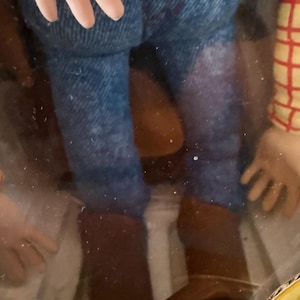 Puede incluir: Figura de acci&oacute;n de Woody, con pantalones vaqueros azules, botas de vaquero marrones y una camisa a cuadros. La figura tiene un cintur&oacute;n marr&oacute;n con una hebilla dorada, parcialmente visible en su embalaje.