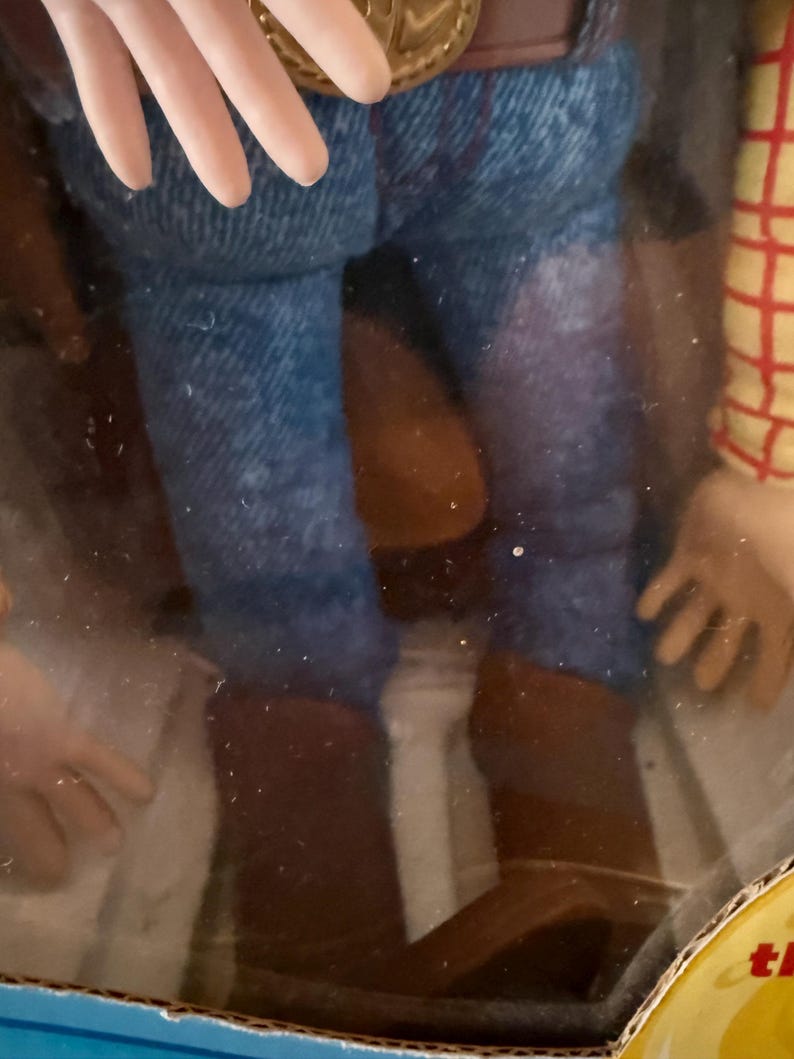 Puede incluir: Figura de acci&oacute;n de Woody, con pantalones vaqueros azules, botas de vaquero marrones y una camisa a cuadros. La figura tiene un cintur&oacute;n marr&oacute;n con una hebilla dorada, parcialmente visible en su embalaje.