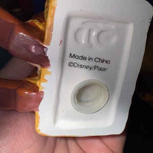 May include: Close-up of the bottom of a ceramic figurine. The base is white with a circular opening and the text "Made in China" and "&copy;Disney/Pixar". The figurine has brown and yellow accents.