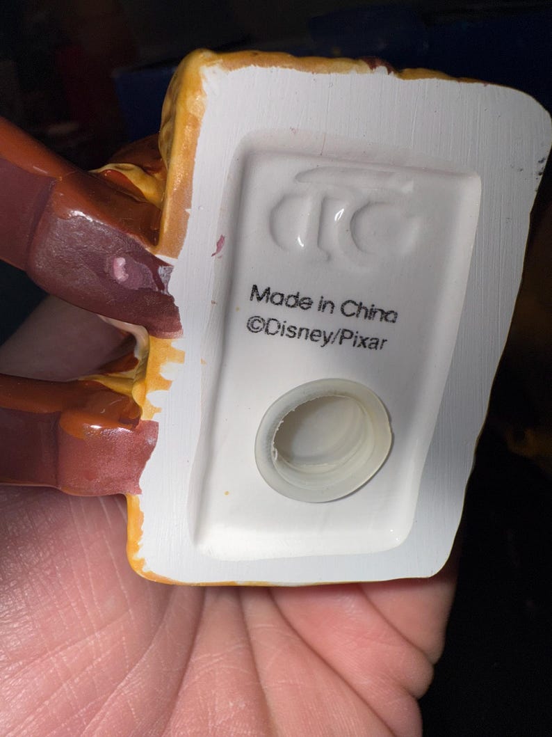 May include: Close-up of the bottom of a ceramic figurine. The base is white with a circular opening and the text "Made in China" and "&copy;Disney/Pixar". The figurine has brown and yellow accents.