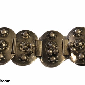 May include: A silver bracelet with a series of oval-shaped links, each with a decorative design of small circles and a flower-like pattern. The links are connected by hinges, allowing the bracelet to be flexible.