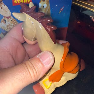 May include: A tan and brown toy horse figurine with an orange saddle and red hooves. The figurine is from the Disney Pixar Toy Story movie. The packaging in the background features Woody and the Toy Story logo.