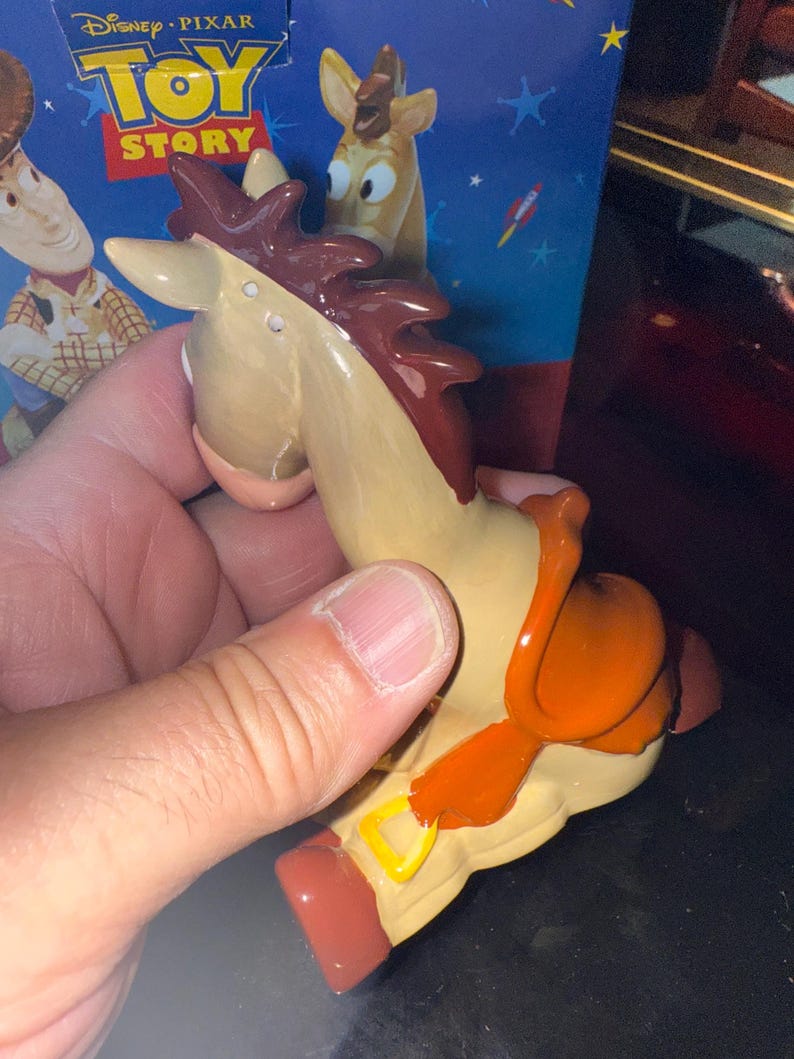 May include: A tan and brown toy horse figurine with an orange saddle and red hooves. The figurine is from the Disney Pixar Toy Story movie. The packaging in the background features Woody and the Toy Story logo.
