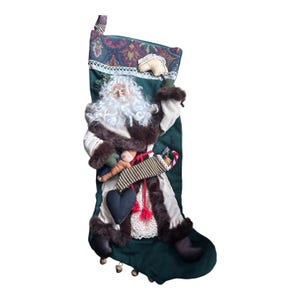 May include: A dark green Christmas stocking featuring a fabric Santa Claus applique with a white faux fur beard and trim. The stocking has a patterned cuff, a small black heart, a striped pouch, and several small metal bells along the bottom edge.
