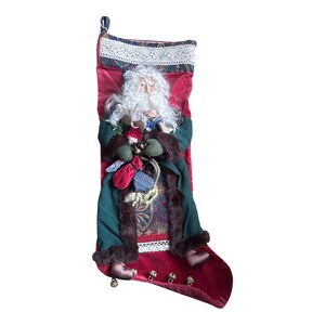 May include: A red velvet Christmas stocking featuring a decorative lace trim at the top and a plush Santa Claus figure attached to the front. The Santa wears a dark green coat with brown faux fur trim and holds small accessories including a red bag.
