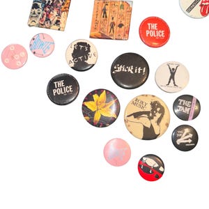 May include: A collection of vintage pinback buttons featuring band logos, album art, and text. The buttons vary in size and shape, with a mix of round and square designs. Colors include red, black, white, pink, and yellow. Some buttons display the text "THE POLICE" and "ROXY MUSIC".