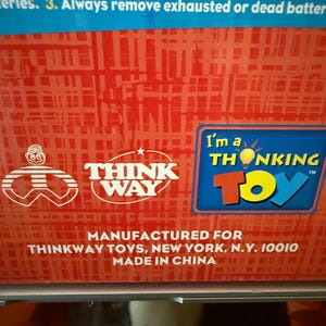 Puede incluir: Primer plano de una caja roja y blanca con el logotipo de Think Way Toys y el texto "I'm a THONKING TOY". La caja tambi&eacute;n incluye instrucciones sobre fugas de bater&iacute;a y el texto "MADE IN CHINA".