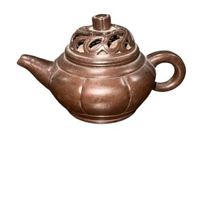 Puede incluir: Una tetera de cerámica marrón con una tapa decorativa con un diseño de celosía. La tetera tiene un cuerpo redondeado y un pico en un lado y un asa en el otro.