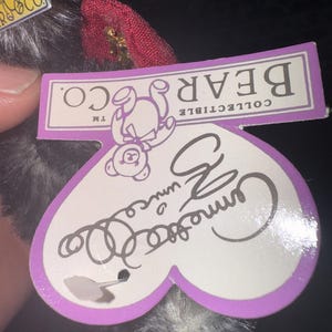 May include: A collectible bear tag with the words "BEAR CO." and "COLLECTIBLE" printed on a purple rectangle. The tag is heart-shaped with the name "Camille" written on it. The bear is black and white.