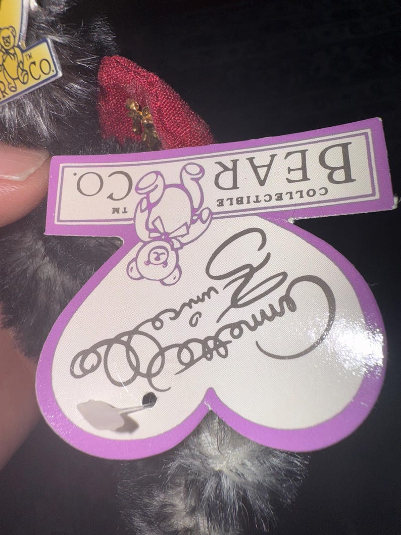 May include: A collectible bear tag with the words "BEAR CO." and "COLLECTIBLE" printed on a purple rectangle. The tag is heart-shaped with the name "Camille" written on it. The bear is black and white.