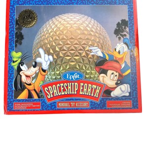 May include: A red box featuring the Epcot Spaceship Earth attraction with Goofy, Mickey Mouse, and Donald Duck. The box reads "Spaceship Earth Monorail Toy Accessory" in a red banner. The background shows the iconic geodesic sphere.