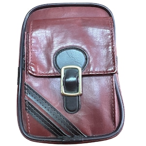 May include: A burgundy leather pouch with a black flap and a gold buckle. The pouch has a black and white striped design on the bottom.