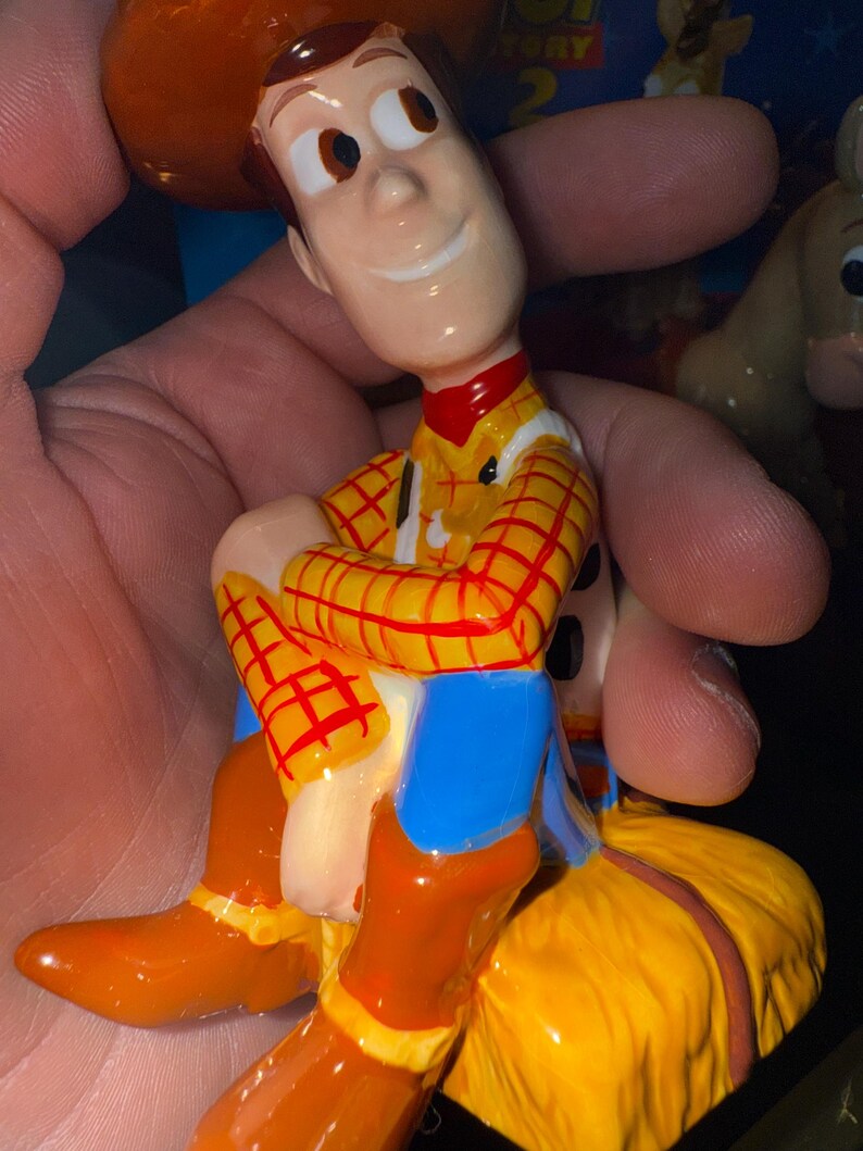 May include: A figurine of the character Woody from the animated film Toy Story. Woody is depicted in his signature cowboy outfit, including a yellow plaid shirt, blue pants, brown boots, and a brown hat. The figurine is held in a hand.